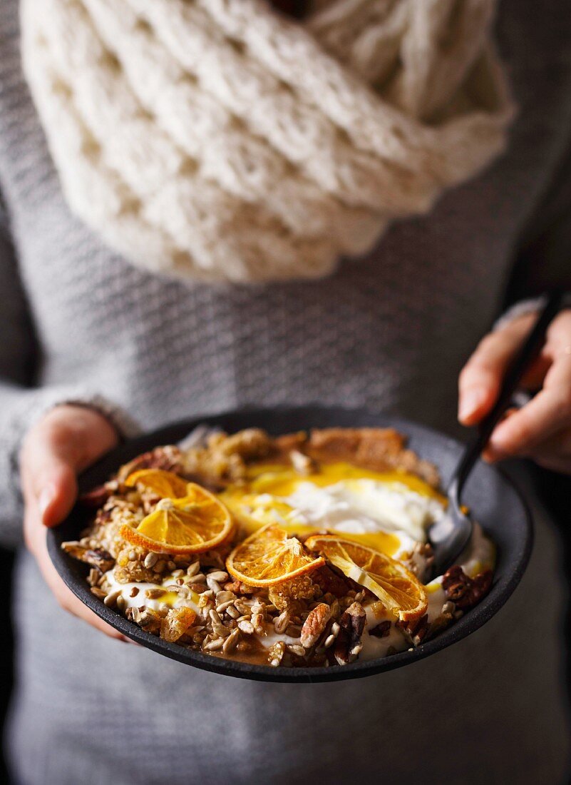 Hand hält Orangen-Smoothie Bowl mit Knuspermüsli und Kokosbutter