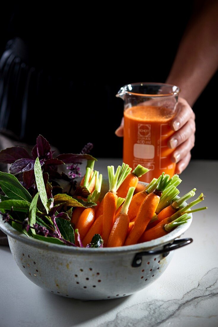 Fresh carrots and herbs in a colander, with carrot juice in measuring cup