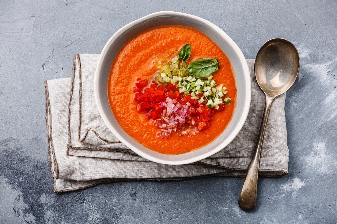Gazpacho soup served with basil and chopped vegetables on grey concrete background