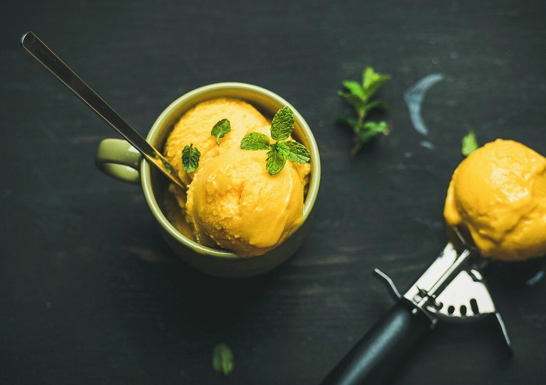 Mango sorbet ice cream scoops with fresh mint in green cup over black wooden background