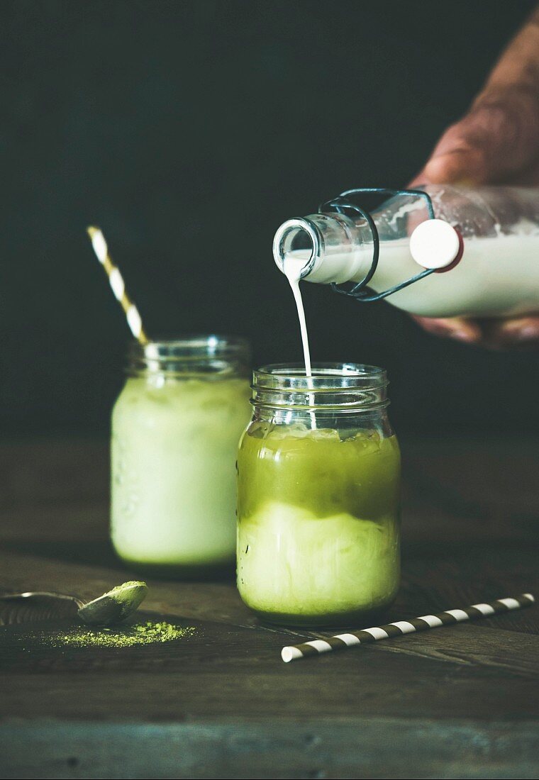 Cold refreshing iced coconut matcha latte drink, hand pouring milk from bottle