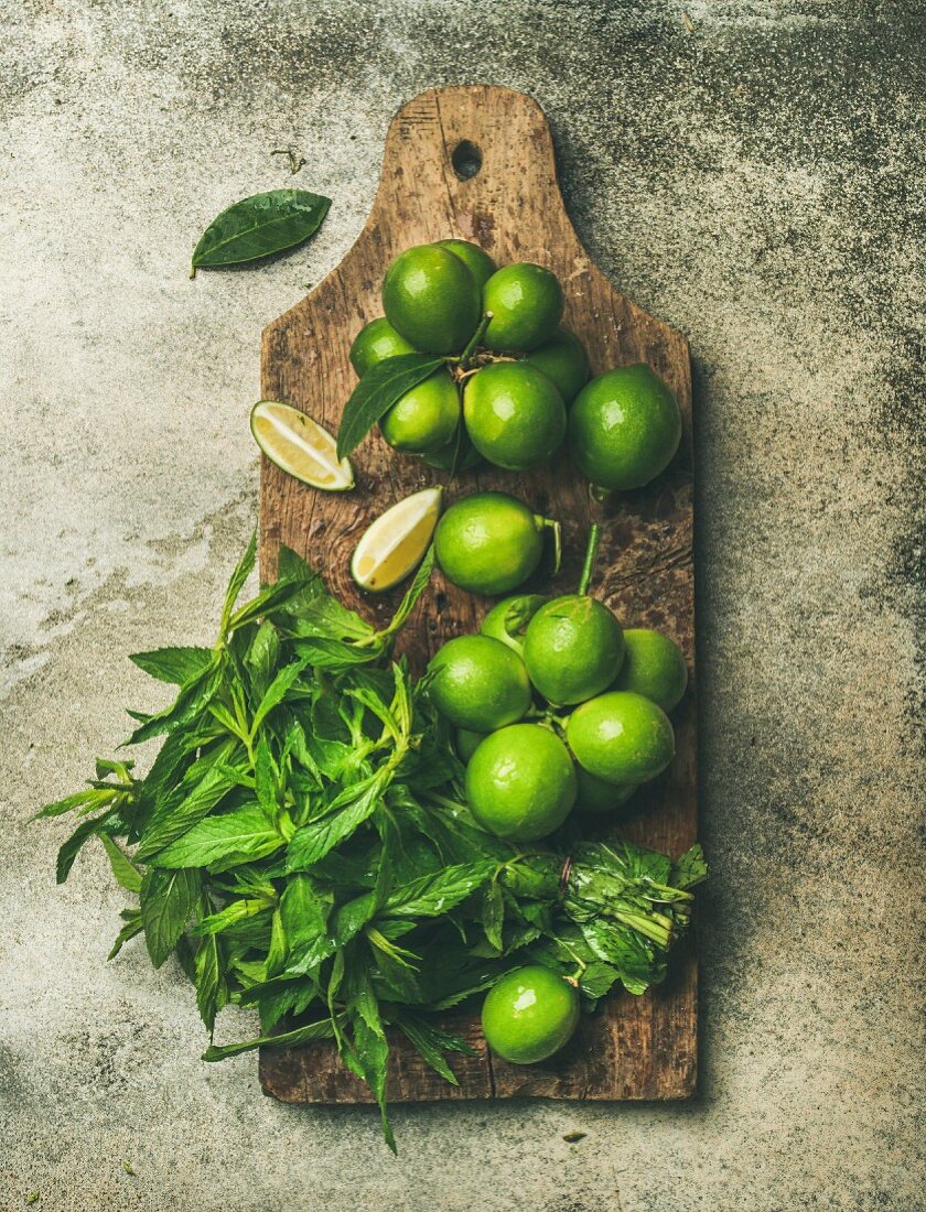 Flatlay of freshly picked organic limes and mint leaves for making cocktail or lemonade on wooden rustic board over grey concrete stone background