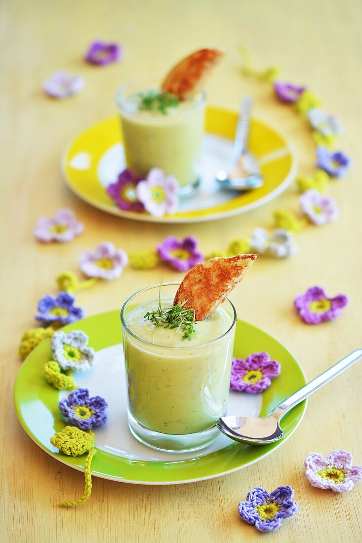Avocadosuppe in zwei Gläsern mit getoasteten Brötchen und frischer Kresse, Frühlingsdekoration aus gehäkelten Blumen