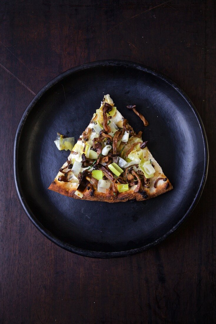 A slice of pizza with mushrooms and leek on a black plate (seen from above)