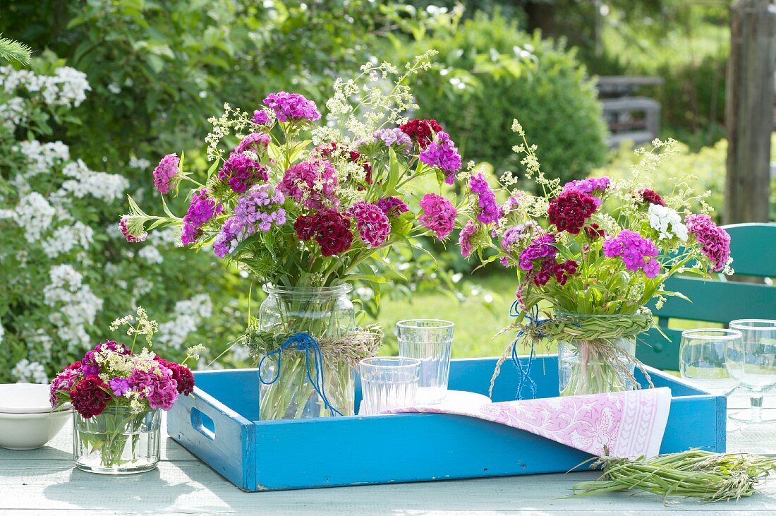 Bouquets of Dianthus barbatus (sweet William) and Galium verum (lady's bedstraw) with plaits of glass tied around jars on blue wooden tray