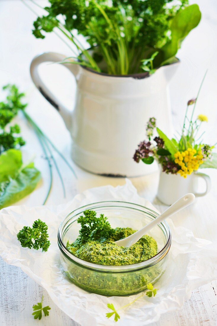 Mixed herb pesto in a glass bowl, with a jug of herbs in the background