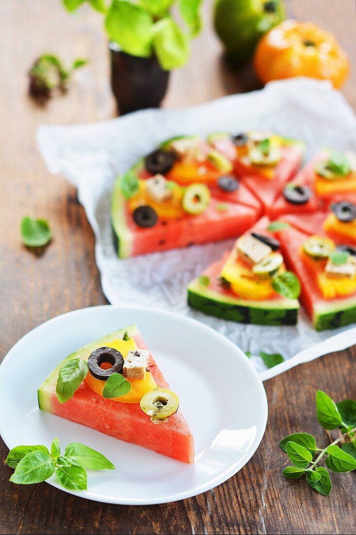 A slice of melon garnished like a pizza with tomatoes, tofu, olives and fresh herbs