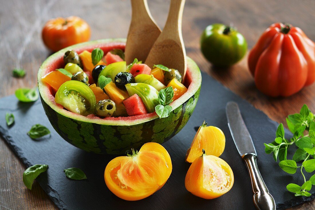 Ausgehöhlte Wassermelone, gefüllt mit Salat aus Wassermelone, bunten Tomaten, Oliven und frischen Kräutern