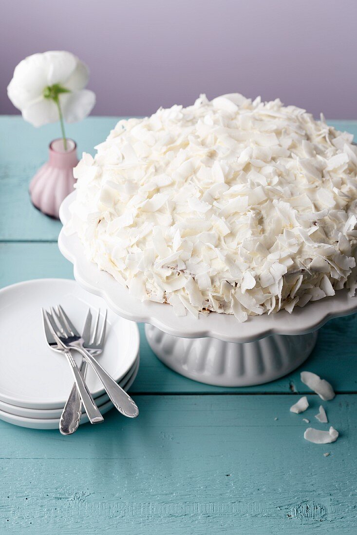 A cake covered with cream cheese and coconut flakes on a white cake stand