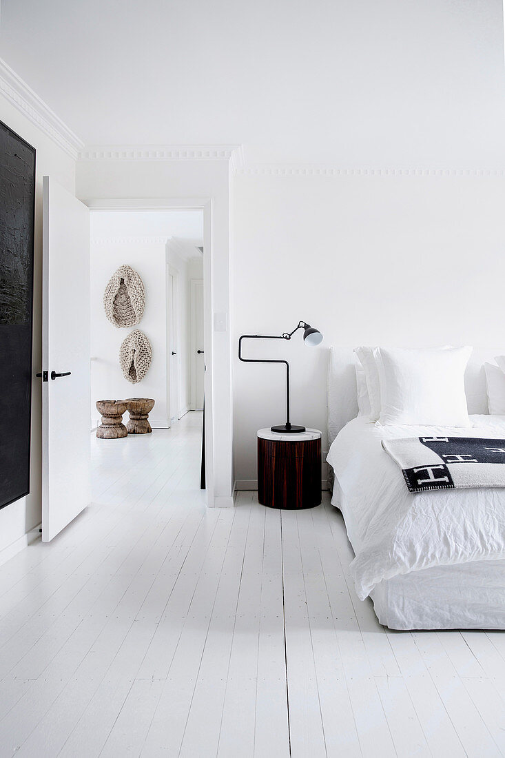 Bedside table with designer lamp next to double bed in bedroom with white floorboards