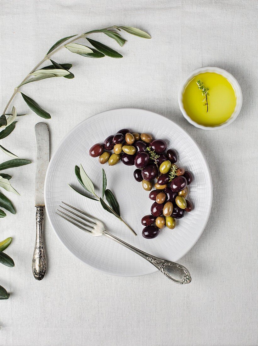 Schwarze und grüne Oliven auf Teller, daneben Besteck und Schälchen mit Olivenöl