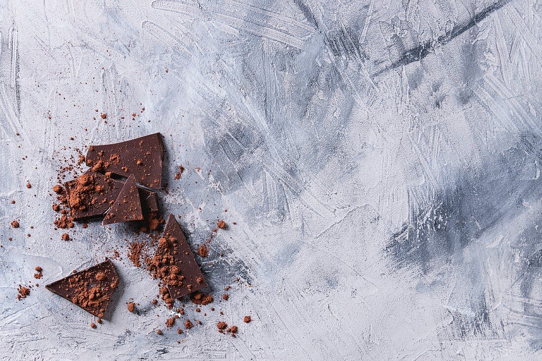 Grob gehackte dunkle Schokolade bestreut mit Kakaopulver auf grauem Untergrund