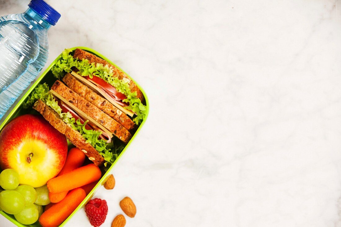 Healthy school lunch box with sandwich, apple, grape, carrot and bottle of water close up on white wooden background