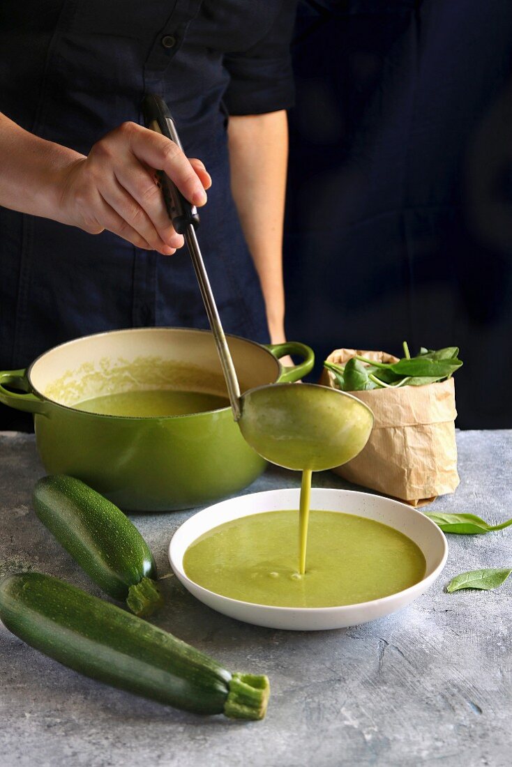 Frau giesst Zucchini-Spinat-Suppe mit Schöpfkelle in Suppenteller
