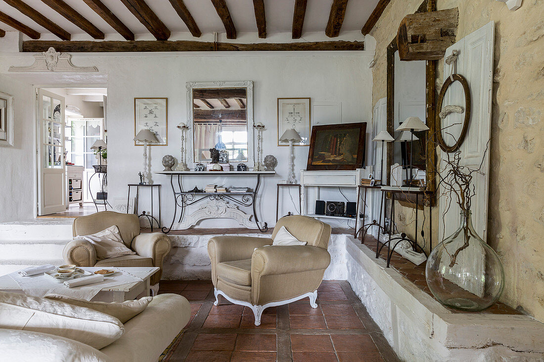 Beige armchair and couch in rustic living room with masonry ledge, terracotta floor tiles and wood-beamed ceiling