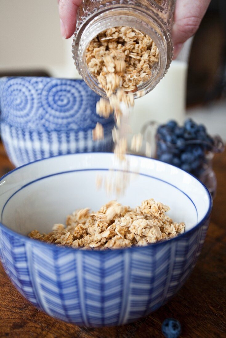 A woman pouring granola from an antique jar into a blue bowl, with blueberries and stacked bowls in the background