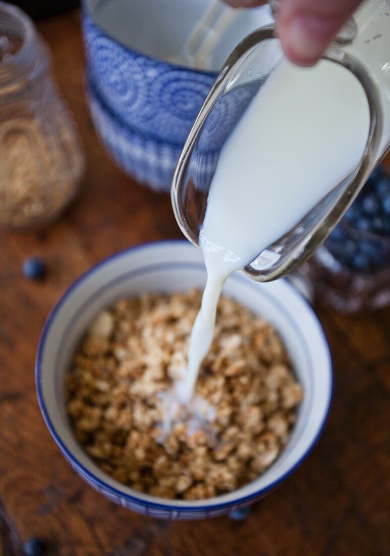 A woman pouring milk from a pitcher onto a bowl of granola, with blueberries, granola and a stack of bowls in the background