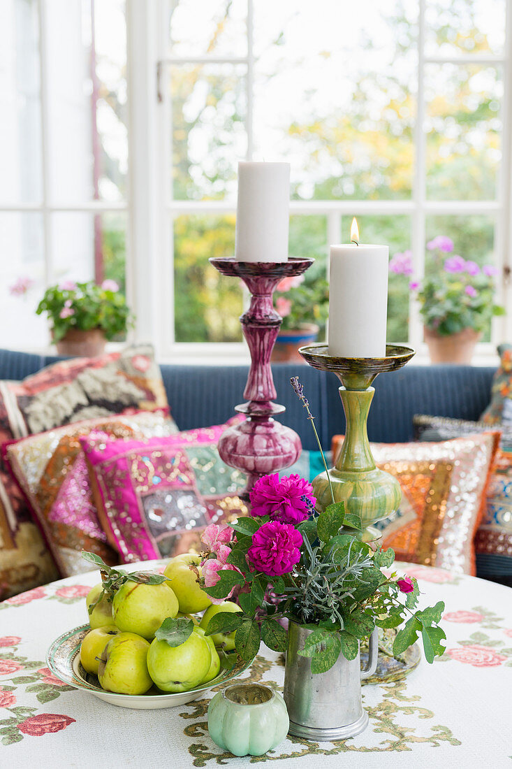 Fruit, flowers and candles on round table in front of sofa with colourful scatter cushions on conservatory