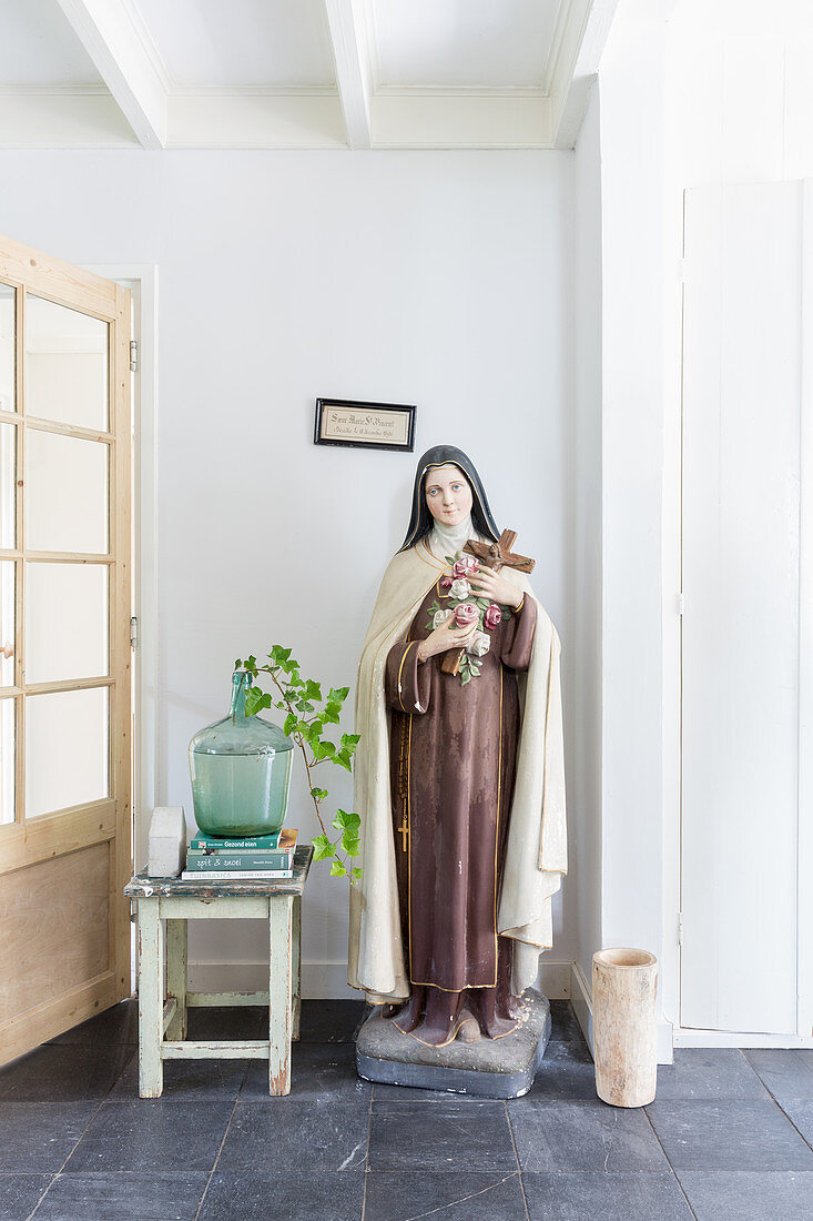 Large statue of the Madonna next to demijohn used as vase on stool