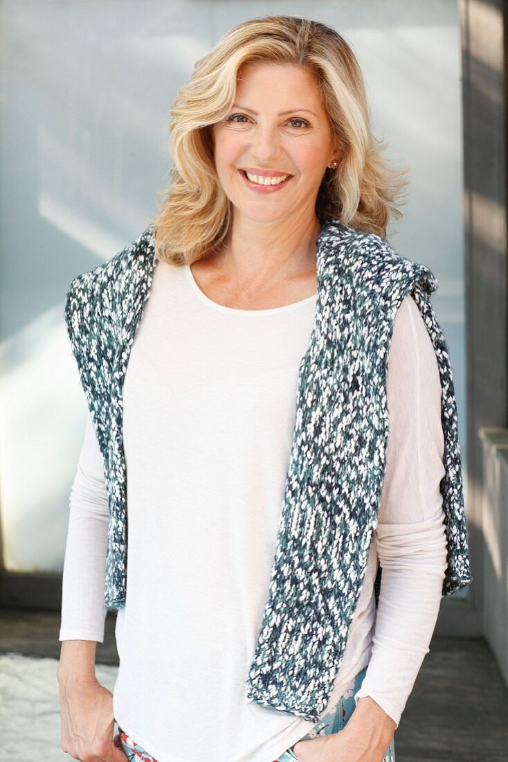 A blonde woman wearing a white, long-sleeved top with a knitted jumper over her shoulders