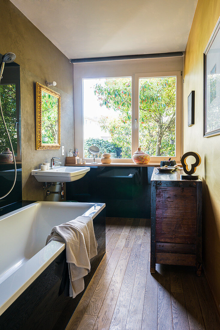 Bathtub, antique sideboard and sink in bathroom with large window and wooden floor