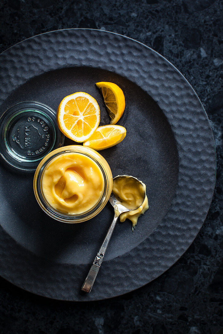 Lemon Curd im Glas auf schwarzem Teller (Draufsicht)