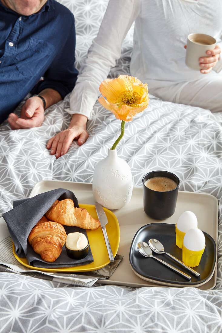 Croissants, eggs, coffee and yellow poppy on breakfast tray on bed with couple in background
