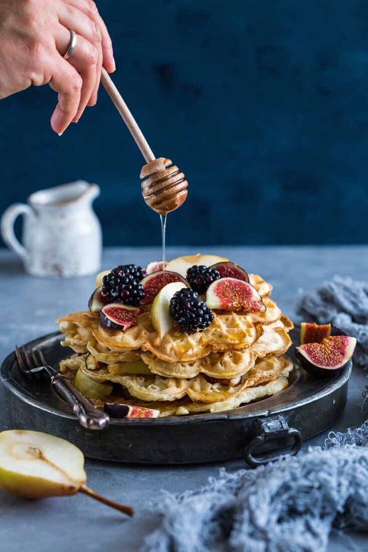 Honey being drizzled over a stack of waffles with berries and figs