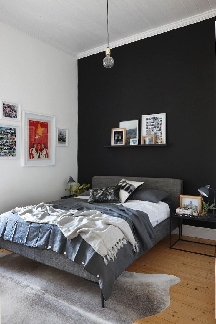 Bed with grey upholstered frame below pictures on narrow shelf on black accent wall
