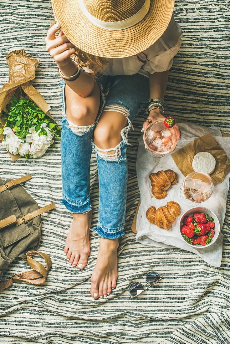 Frau in Jeans mit Weinglas, Erdbeeren und Croissants auf Picknickdecke