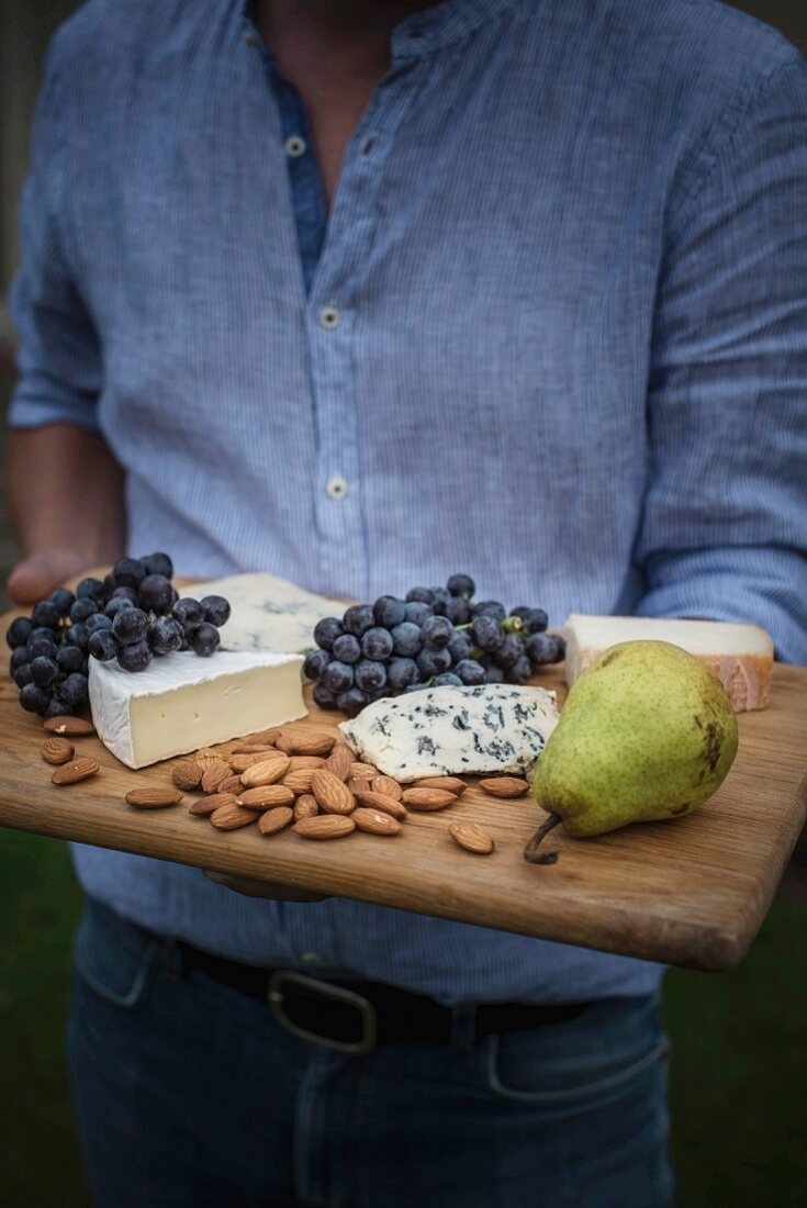 A man holding a cheese board with grapes, pear and almonds