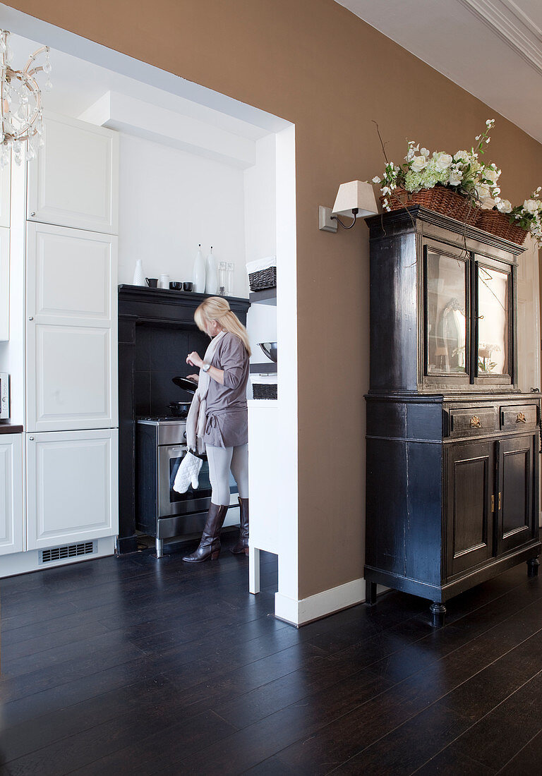 Antique sideboard with basket of flowers against light brown wall next to open doorway: woman in kitchen in background