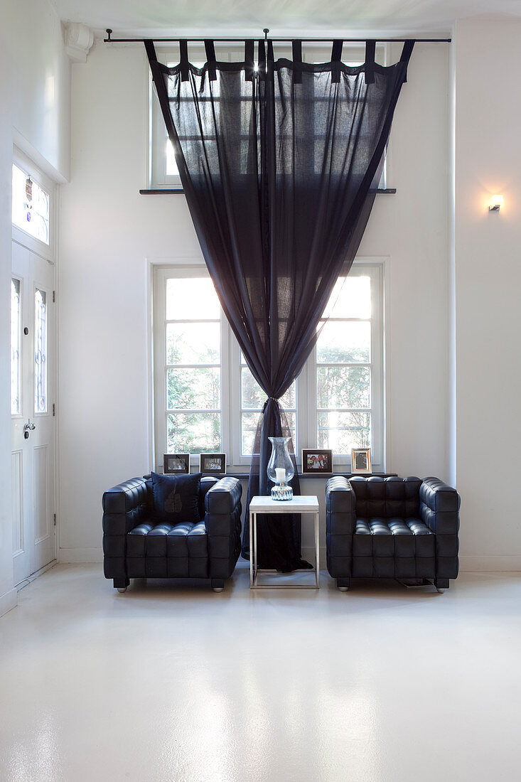 Two black leather armchairs in room with high ceiling and dark curtains on windows