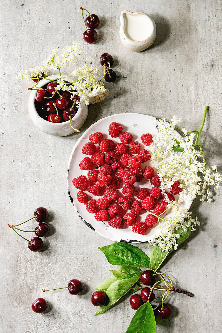 Fresh cherry and raspberry berries in ceramic mug and plate, elderflowers, jug of cream