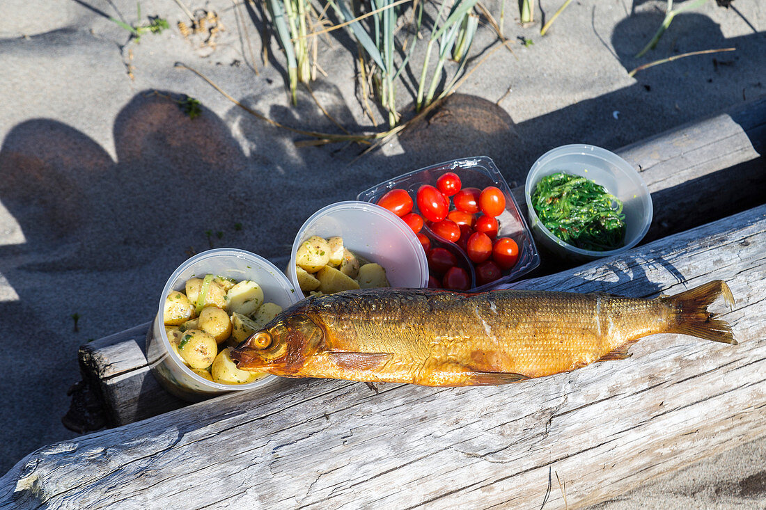 Geräucherter Fisch, Kartoffeln, Tomaten und Kräuter auf Holzsteg am Strand