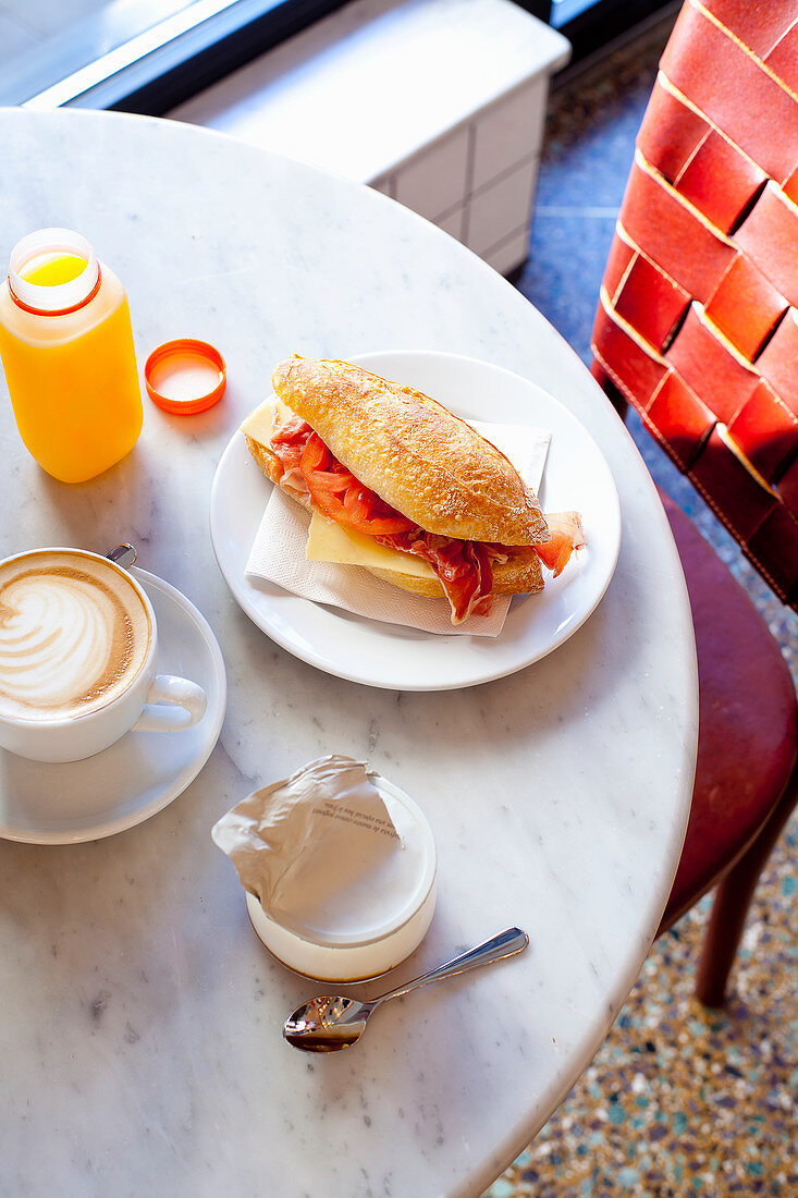 Breakfast consisting of a sandwich, coffee, orange juice and yoghurt on a bistro table