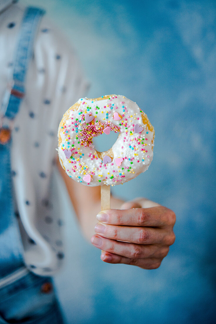 Person hält Donut am Stiel dekoriert mit Zuckerglasur und bunten Zuckerperlen