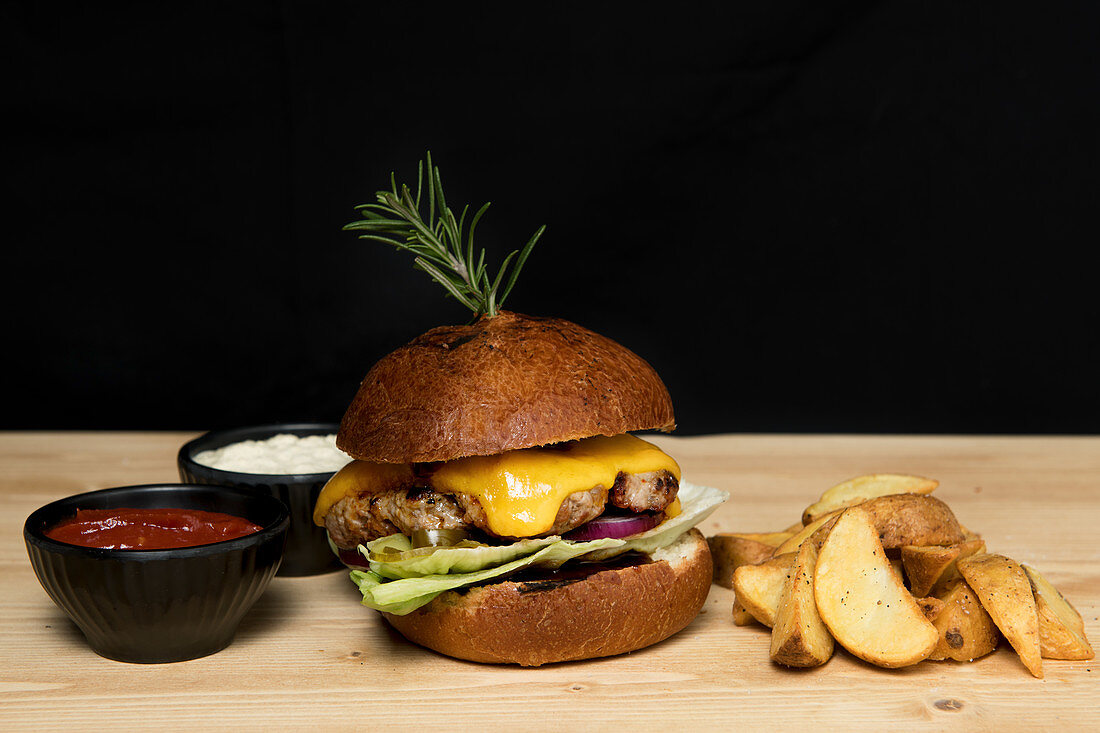 Hamburger mit Kartoffelspalten und zweierlei Dips vor schwarzem Hintergrund