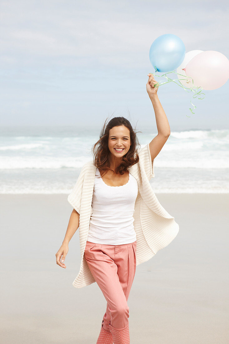 Brünette Frau mit Luftballons in Strickjacke und rosa Hose am Meer