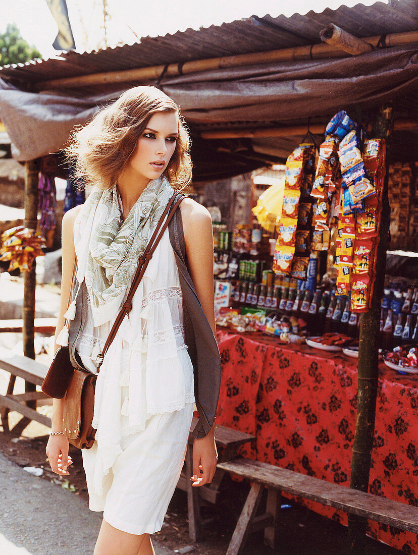 A young blonde woman wearing a white shirt and shorts with a scarf and a waistcoat with a shoulder bag