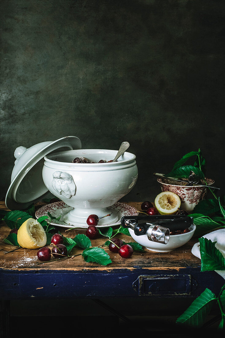 Composition of ceramic bowl with cherry and sugar and cherry kernel remover on table with green leaves