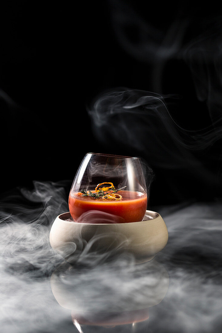 Composition of glass in bowl filled with red spicy alcohol cocktail and served on table in smoke against black background