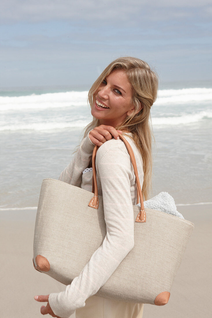 A blond woman on a beach wearing a light cardigan and shorts and holding a bag