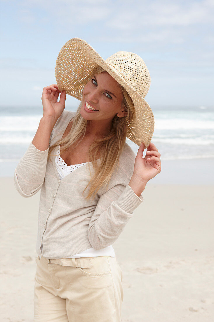 Blonde Frau mit Sommerhut in hellem Cardigan und Shorts am Strand