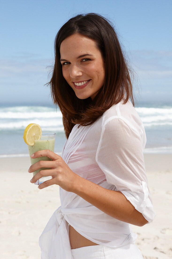 Junge brünette Frau mit Smoothie in weißem Hemd am Strand