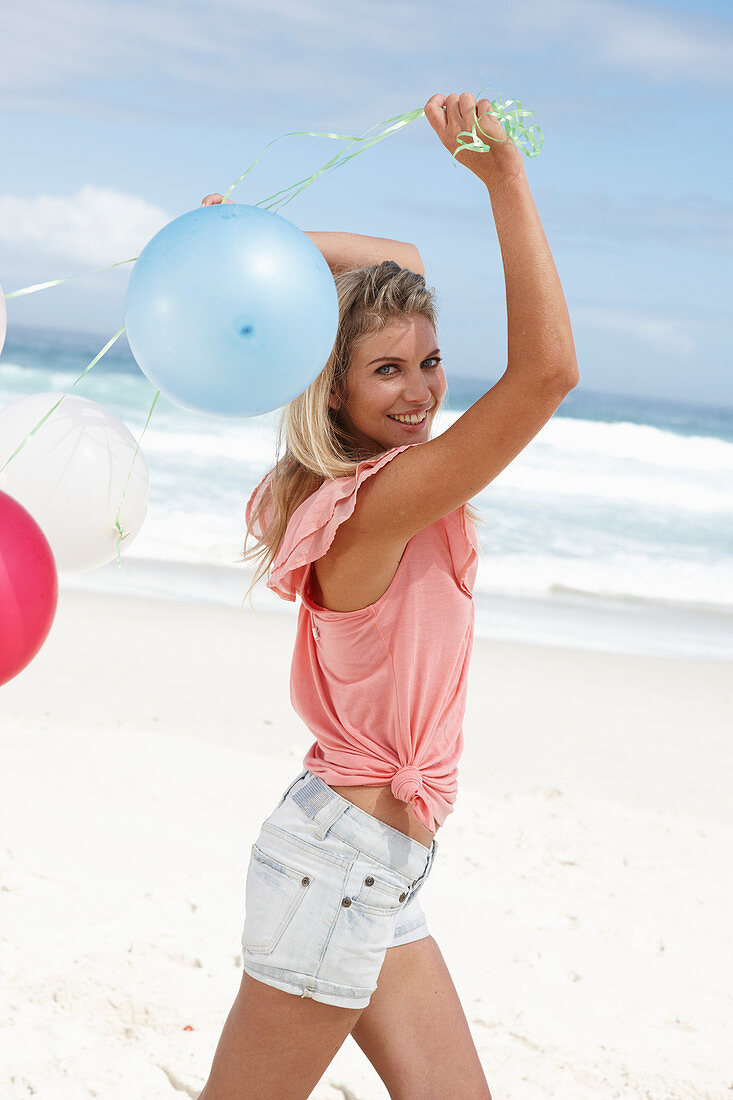 Junge Frau mit Luftballons im rosa Top und Jeansshorts am Strand