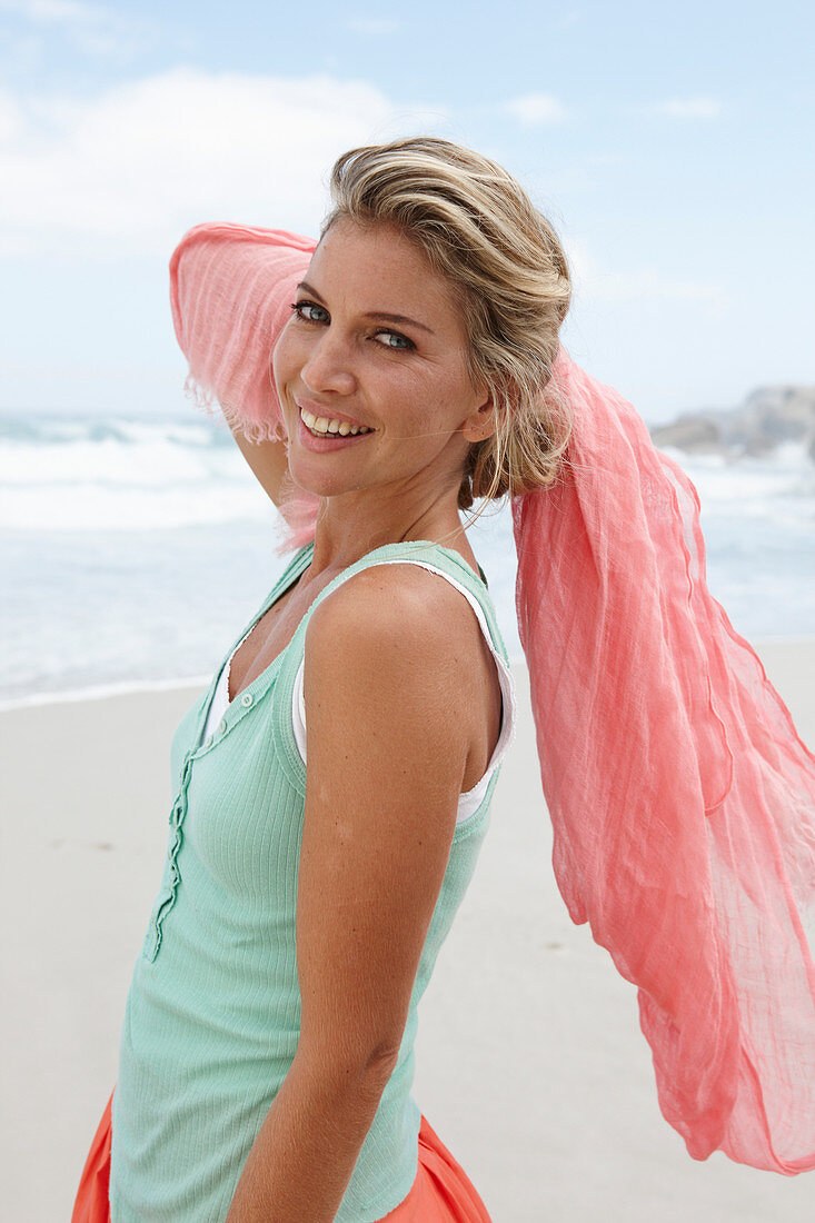 A blonde woman on a beach with a scarf wearing a turquoise top and a salmon-pink skirt