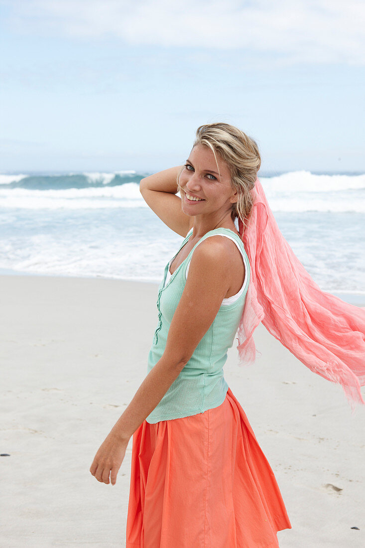 Blonde Frau mit Tuch in türkisgrünem Top und lachsfarbenem Rock am Strand