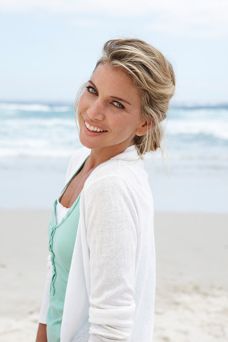 Blonde Frau in türkisgrünem Top und weißer Strickjacke am Strand