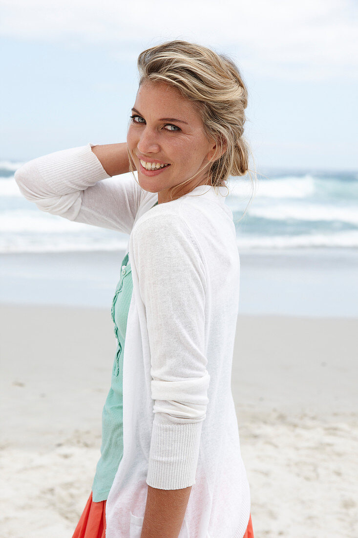 A blonde woman on a beach wearing a turquoise top and a white cardigan